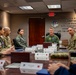 A military briefing or meeting is taking place in a formal conference room, with personnel in various U.S. Air Force uniforms seated around a large reflective table. A senior officer in a green flight suit appears to be leading the discussion from the head of the table. Name placards are visible in front of attendees, and a unit crest with a partially visible motivational quote is displayed on the wall behind the leader.