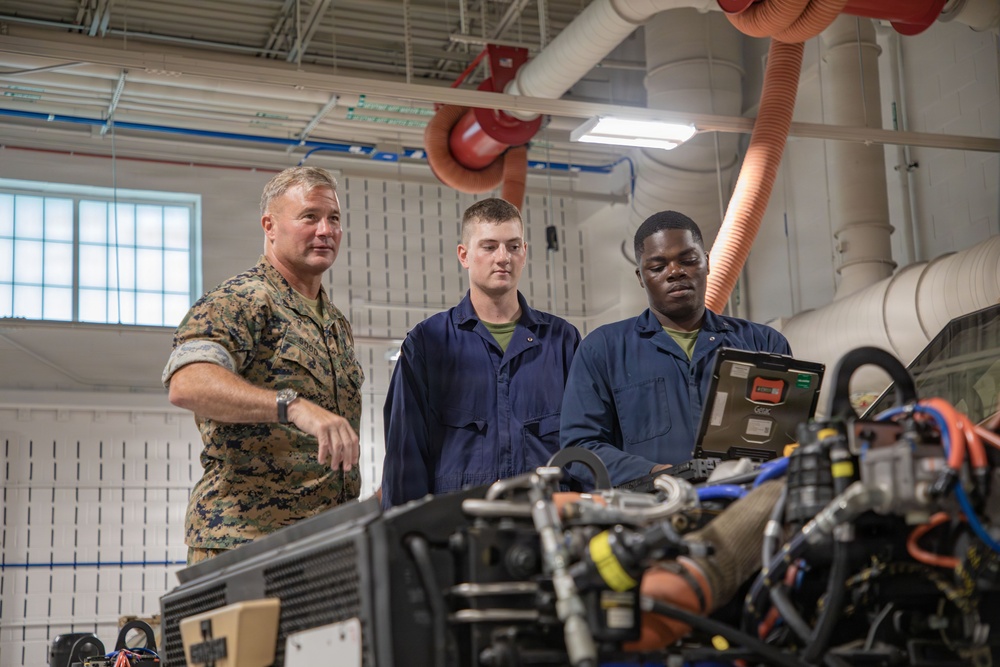 Three military personnel are gathered around an engine or mechanical system in what appears to be a maintenance bay or motor pool facility. A senior Marine in camouflage utilities is observing or instructing two junior personnel wearing blue mechanic coveralls who are using a ruggedized laptop for diagnostics. The setting suggests a vehicle or equipment maintenance training or inspection scenario.