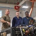 Three military personnel are gathered around an engine or mechanical system in what appears to be a maintenance bay or motor pool facility. A senior Marine in camouflage utilities is observing or instructing two junior personnel wearing blue mechanic coveralls who are using a ruggedized laptop for diagnostics. The setting suggests a vehicle or equipment maintenance training or inspection scenario.