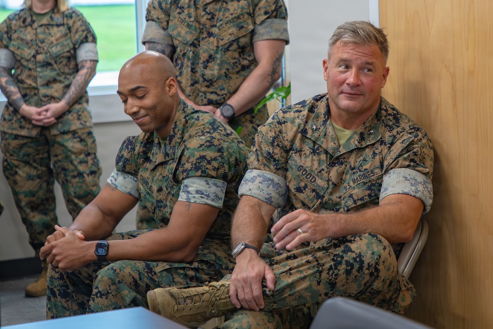 The image shows several U.S. Marines in MARPAT (Marine Pattern) camouflage uniforms seated and standing in what appears to be an indoor facility or briefing room. Two Marines are prominently seated in the foreground, with the one on the right bearing a name tape reading 'GOOD' and a rank insignia consistent with a general officer (one star visible). The setting suggests an official visit, briefing, or meeting environment.