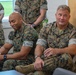 The image shows several U.S. Marines in MARPAT (Marine Pattern) camouflage uniforms seated and standing in what appears to be an indoor facility or briefing room. Two Marines are prominently seated in the foreground, with the one on the right bearing a name tape reading 'GOOD' and a rank insignia consistent with a general officer (one star visible). The setting suggests an official visit, briefing, or meeting environment.