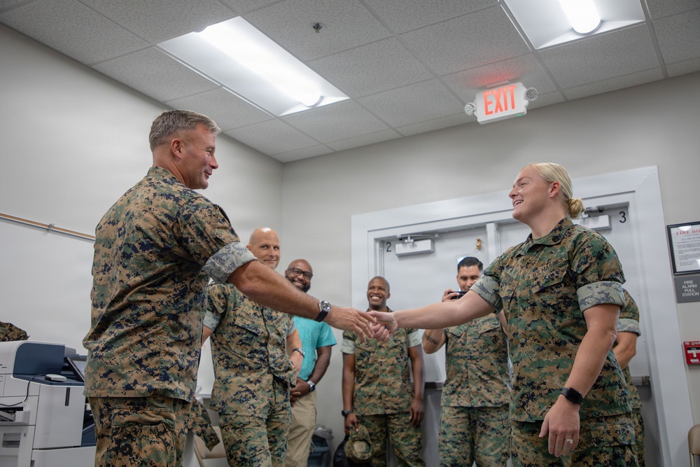 Two U.S. Marines in MARPAT camouflage uniforms are shaking hands in what appears to be an indoor military facility or office space. Several onlookers, including both uniformed personnel and civilians, are smiling in the background. The scene suggests a formal recognition, greeting, or award ceremony.