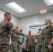 Two U.S. Marines in MARPAT camouflage uniforms are shaking hands in what appears to be an indoor military facility or office space. Several onlookers, including both uniformed personnel and civilians, are smiling in the background. The scene suggests a formal recognition, greeting, or award ceremony.