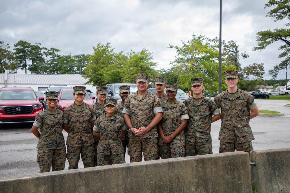 A group of approximately ten U.S. Marine Corps personnel in MARPAT woodland camouflage uniforms pose together for a group photo in a parking lot. The group includes both male and female Marines of varying ranks, gathered around what appears to be a senior non-commissioned or commissioned officer in the center. The setting appears to be a military installation, likely during overcast weather conditions.