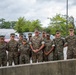 A group of approximately ten U.S. Marine Corps personnel in MARPAT woodland camouflage uniforms pose together for a group photo in a parking lot. The group includes both male and female Marines of varying ranks, gathered around what appears to be a senior non-commissioned or commissioned officer in the center. The setting appears to be a military installation, likely during overcast weather conditions.