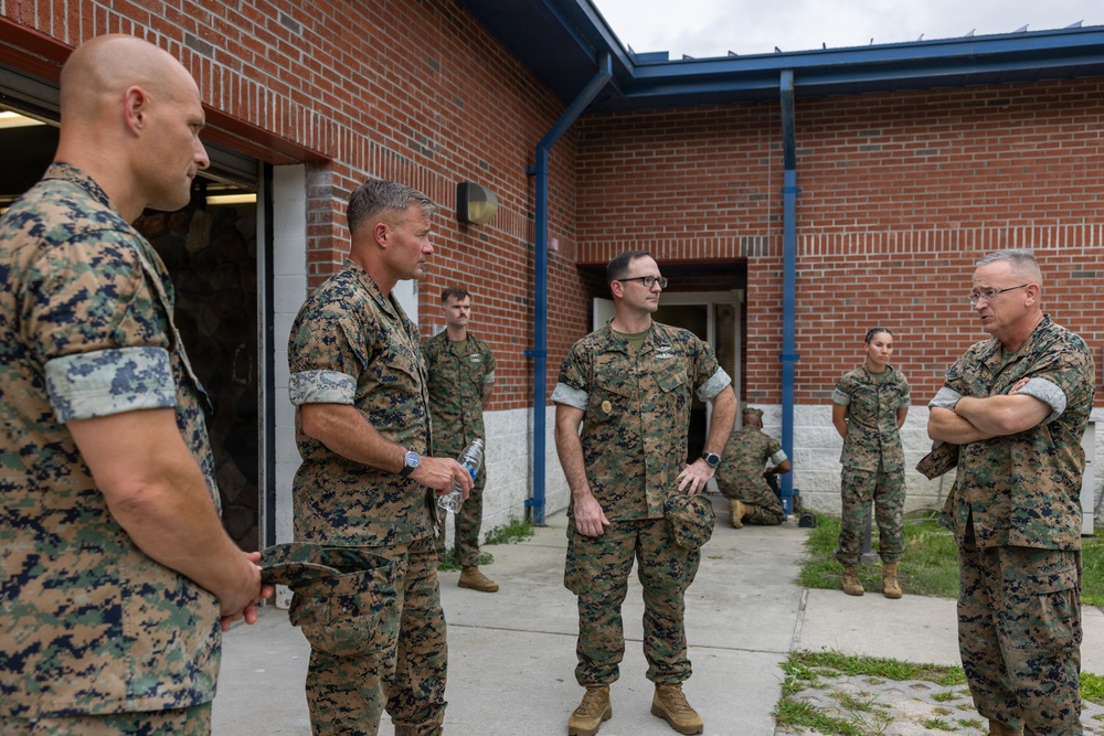 A group of U.S. Marines in MARPAT woodland camouflage uniforms are gathered outside a brick building, engaged in what appears to be a briefing or informal discussion. Several personnel of varying ranks are standing in a courtyard area, with one senior figure on the right standing with arms crossed. The setting appears to be a military installation or base facility.