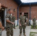 A group of U.S. Marines in MARPAT woodland camouflage uniforms are gathered outside a brick building, engaged in what appears to be a briefing or informal discussion. Several personnel of varying ranks are standing in a courtyard area, with one senior figure on the right standing with arms crossed. The setting appears to be a military installation or base facility.