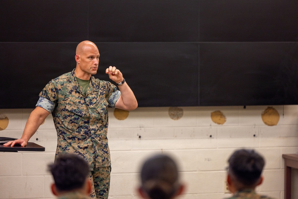 A U.S. Marine in MARPAT digital camouflage uniform is delivering a briefing or lecture to a group of seated military personnel. He is standing at the front of what appears to be a classroom or training room, gesturing with his right hand in a fist to emphasize a point. The setting suggests a formal military instruction or leadership engagement session.