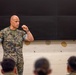 A U.S. Marine in MARPAT digital camouflage uniform is delivering a briefing or lecture to a group of seated military personnel. He is standing at the front of what appears to be a classroom or training room, gesturing with his right hand in a fist to emphasize a point. The setting suggests a formal military instruction or leadership engagement session.