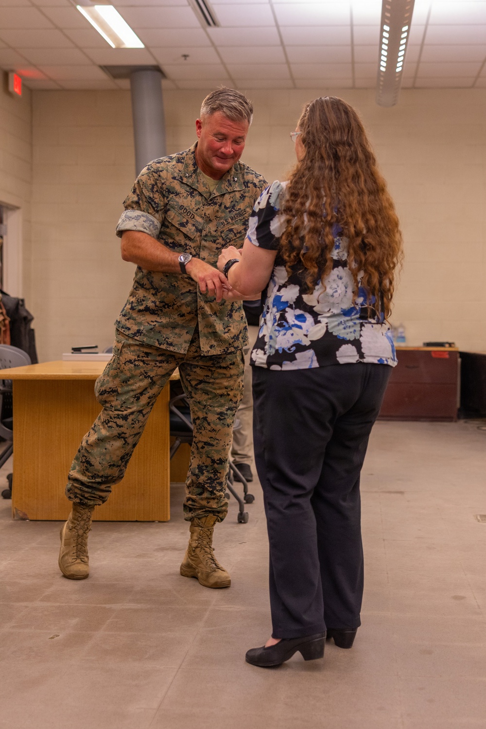 A U.S. Marine in MARPAT camouflage uniform, with the name tape 'GOOD' visible, interacts with a civilian woman in a floral blouse inside what appears to be an office or briefing room. The Marine appears to be examining or demonstrating something on the woman's wrist or arm. The setting is an indoor military or government facility with standard drop-ceiling fluorescent lighting.