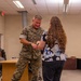 A U.S. Marine in MARPAT camouflage uniform, with the name tape 'GOOD' visible, interacts with a civilian woman in a floral blouse inside what appears to be an office or briefing room. The Marine appears to be examining or demonstrating something on the woman's wrist or arm. The setting is an indoor military or government facility with standard drop-ceiling fluorescent lighting.