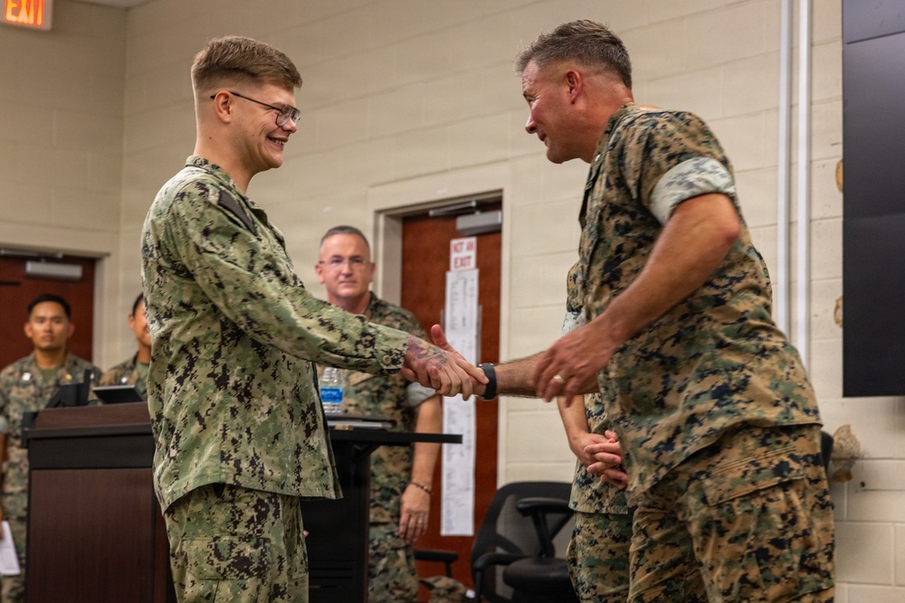 Two military personnel are shaking hands in what appears to be an indoor military facility, likely during a formal recognition or award ceremony. The individual on the left is wearing a U.S. Navy NWU Type I (digital blue/green) uniform, while the individual on the right is wearing a U.S. Marine Corps MARPAT woodland uniform. Both are smiling, suggesting a positive or celebratory interaction.