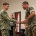 Two military personnel are shaking hands in what appears to be an indoor military facility, likely during a formal recognition or award ceremony. The individual on the left is wearing a U.S. Navy NWU Type I (digital blue/green) uniform, while the individual on the right is wearing a U.S. Marine Corps MARPAT woodland uniform. Both are smiling, suggesting a positive or celebratory interaction.