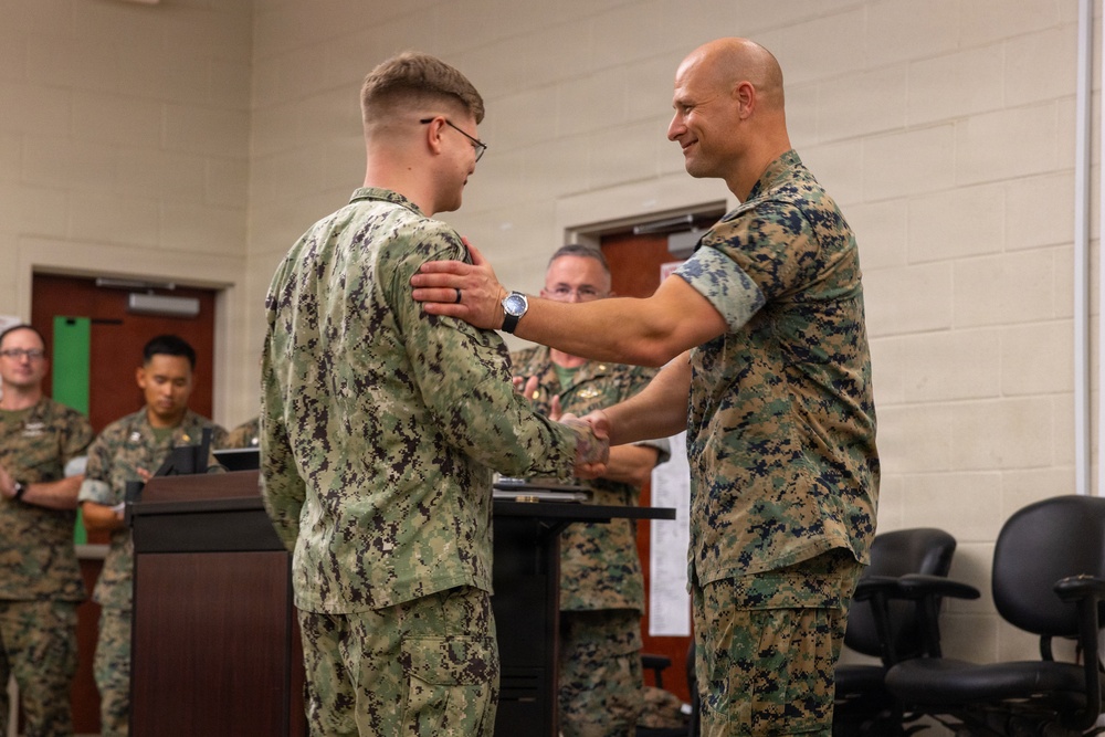 A military ceremony is taking place indoors where a senior officer in Marine Corps MARPAT camouflage is shaking hands with and placing his hand on the shoulder of a junior service member in Navy Working Uniform (NWU) digital camouflage. The gesture suggests an award presentation, promotion, or recognition ceremony. Several other military personnel in various camouflage uniforms are observing in the background near a podium.