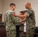 A military ceremony is taking place indoors where a senior officer in Marine Corps MARPAT camouflage is shaking hands with and placing his hand on the shoulder of a junior service member in Navy Working Uniform (NWU) digital camouflage. The gesture suggests an award presentation, promotion, or recognition ceremony. Several other military personnel in various camouflage uniforms are observing in the background near a podium.