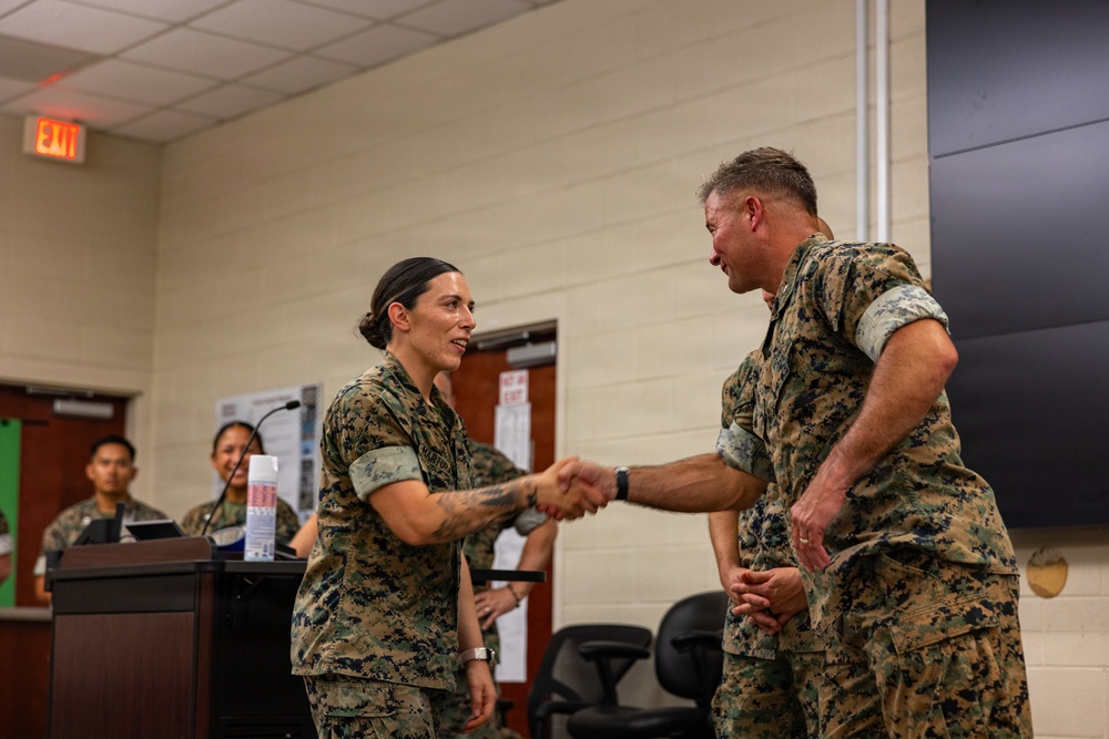 Two U.S. Marines in MARPAT camouflage uniforms are shaking hands in what appears to be an indoor military briefing or ceremony room. A female Marine and a male Marine are the central figures, with additional personnel visible at a podium in the background. The setting suggests a formal recognition or award ceremony.