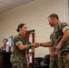 Two U.S. Marines in MARPAT camouflage uniforms are shaking hands in what appears to be an indoor military briefing or ceremony room. A female Marine and a male Marine are the central figures, with additional personnel visible at a podium in the background. The setting suggests a formal recognition or award ceremony.