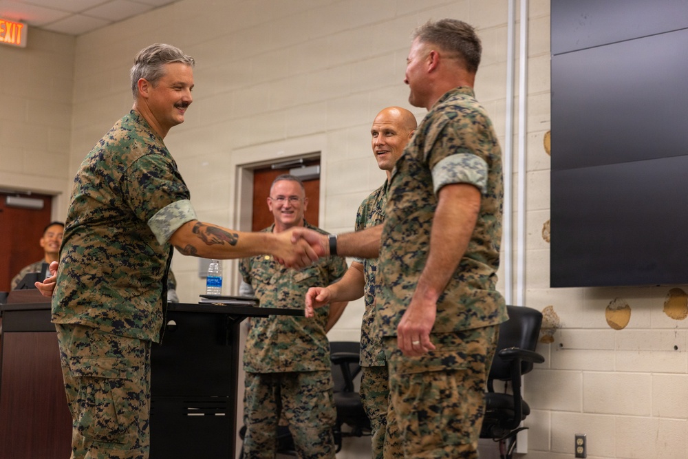 Several military personnel in MARPAT (Marine Pattern) camouflage uniforms are gathered in what appears to be a briefing or conference room. Two individuals in the foreground are shaking hands in what appears to be a formal recognition or greeting ceremony, while two others observe in the background. The setting suggests an official military function such as an award presentation, promotion, or unit ceremony.