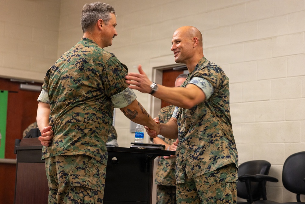 Two military personnel in MARPAT (Marine pattern) camouflage uniforms are shaking hands in what appears to be an indoor briefing or ceremony room. The individual on the right is smiling broadly and extending his other hand in a congratulatory or welcoming gesture. Additional uniformed personnel are visible in the background near a podium.