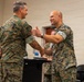 Two military personnel in MARPAT (Marine pattern) camouflage uniforms are shaking hands in what appears to be an indoor briefing or ceremony room. The individual on the right is smiling broadly and extending his other hand in a congratulatory or welcoming gesture. Additional uniformed personnel are visible in the background near a podium.