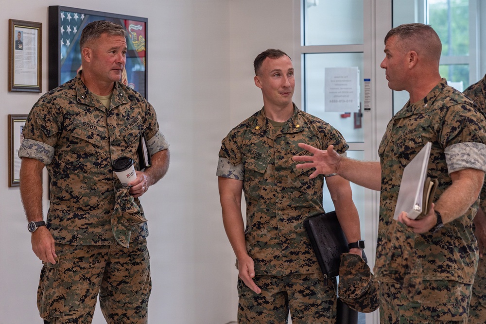 Three U.S. Marine Corps personnel in MARPAT woodland camouflage uniforms are engaged in conversation inside what appears to be a military building or office. The leftmost individual has a name tape reading 'GOOD' and is holding a coffee cup and notebook, while the center individual has a gold oak leaf insignia indicating the rank of Major. They appear to be in a briefing or informal discussion setting.