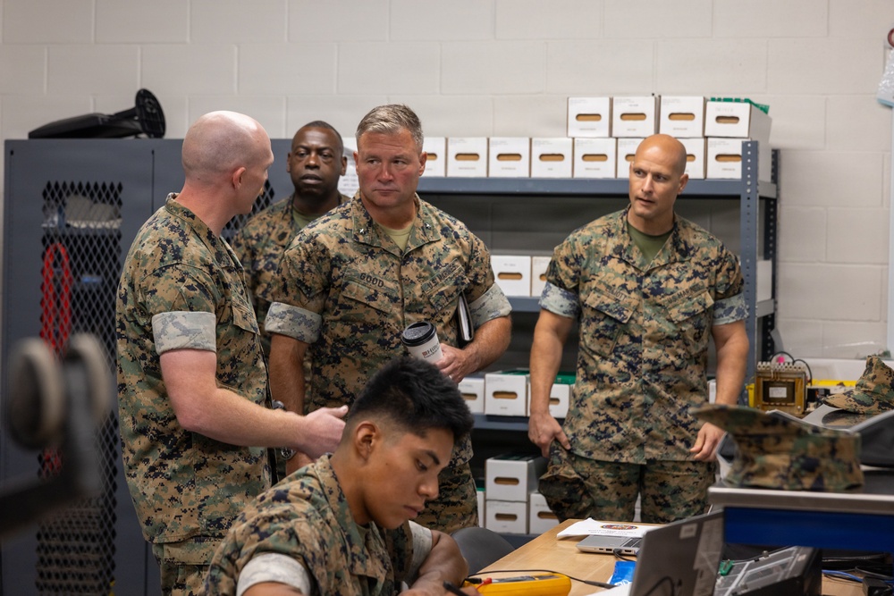 Several U.S. Marine Corps personnel in MARPAT camouflage uniforms are gathered in what appears to be a maintenance or technical workspace. A senior officer with the name tape 'GOOD' and visible rank insignia is observing a junior Marine working at a desk with electronic equipment. The scene appears to be an official visit or inspection of a technical facility.