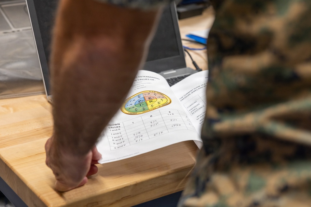 A person in military camouflage uniform is leaning over a wooden desk examining a technical document or textbook. The document displays an Ohm's Law wheel/pie chart diagram along with a reference table showing electrical formulas involving Current (I), Voltage/EMF (E), Resistance (R), and Power (P). A laptop is visible in the background on the same desk.