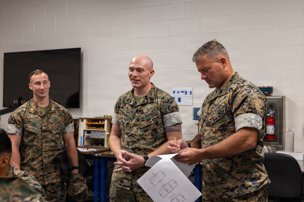 Three U.S. Marines in MARPAT camouflage uniforms are gathered in what appears to be a training or workshop facility. A senior Marine on the right is reviewing a document with printed diagrams while holding a pencil, as a middle Marine gestures and appears to be explaining something. A third Marine stands in the background observing the interaction.