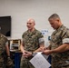 Three U.S. Marines in MARPAT camouflage uniforms are gathered in what appears to be a training or workshop facility. A senior Marine on the right is reviewing a document with printed diagrams while holding a pencil, as a middle Marine gestures and appears to be explaining something. A third Marine stands in the background observing the interaction.