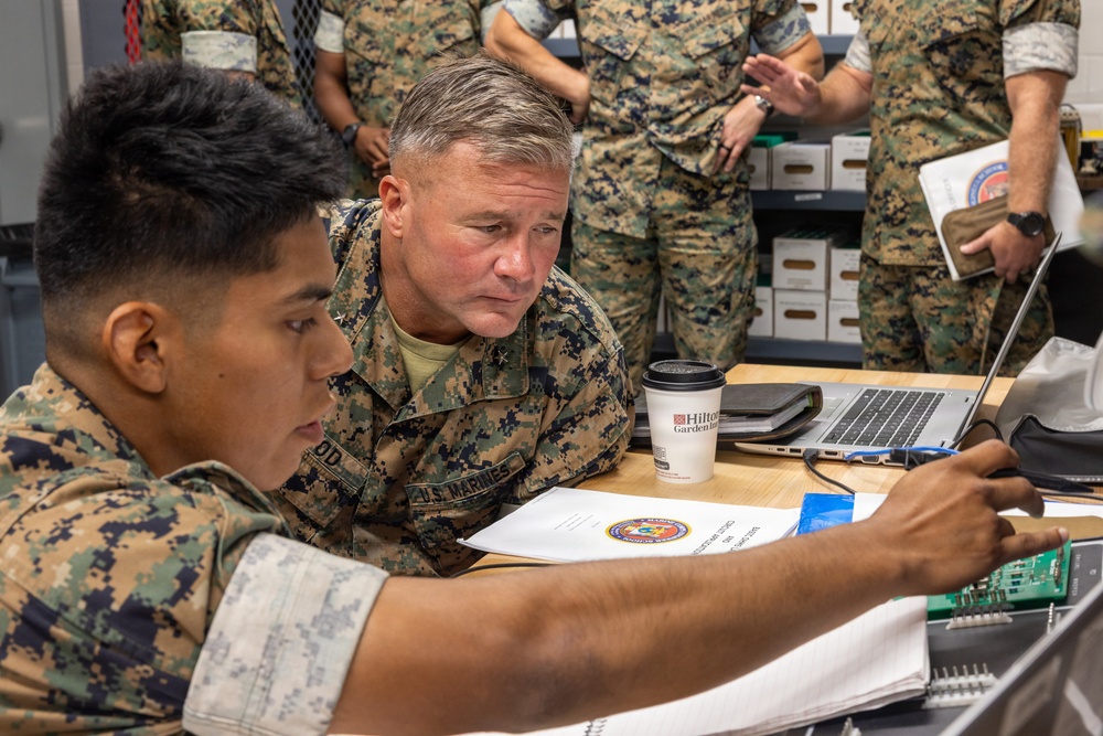 Two U.S. Marines in MARPAT digital camouflage uniforms are examining electronic equipment at a workstation. A senior officer leans in to observe what appears to be a circuit board or electronic component being handled by a junior Marine. Several other Marines are visible in the background of what appears to be an office or operations room.