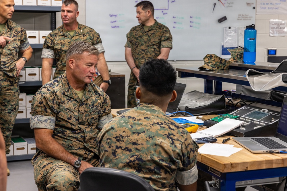 A group of U.S. Marines in MARPAT camouflage uniforms are gathered in what appears to be a technical workspace or classroom setting. A senior Marine, identified by his name tape as 'GOOD,' is seated and engaged in conversation with a junior Marine seated at a workbench with electronic equipment and a laptop. Several other Marines stand in the background observing.