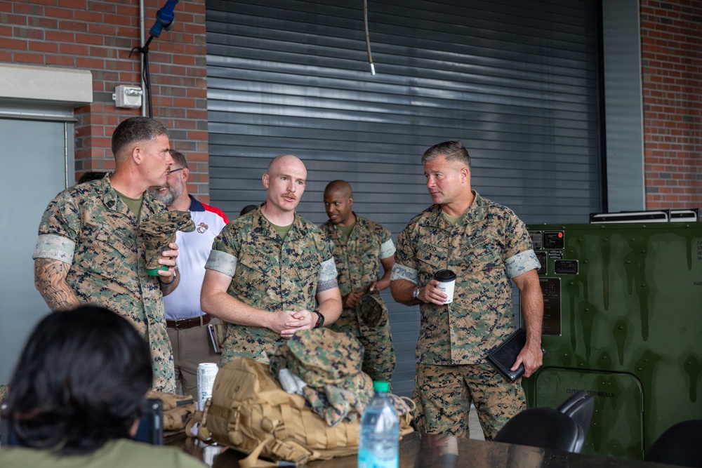 Several U.S. Marines in MARPAT woodland camouflage uniforms are engaged in conversation in what appears to be a hangar or bay facility. Three Marines are prominently featured in the foreground, with one holding a coffee cup and another holding a beverage can. Military equipment and gear are visible on a table in the foreground.