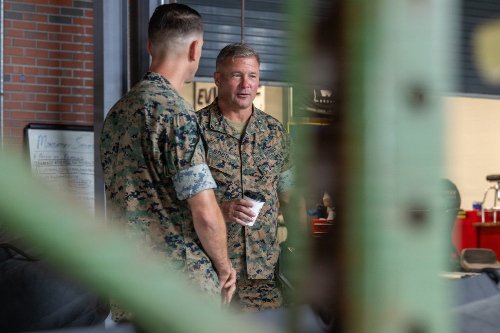 Two U.S. Marines in MARPAT woodland camouflage uniforms are engaged in conversation inside what appears to be a military facility or training building. The Marine facing the camera has the name tape 'GOOD' visible on his uniform and is holding a disposable coffee cup. The image is shot through foreground foliage, creating a partially obscured perspective.