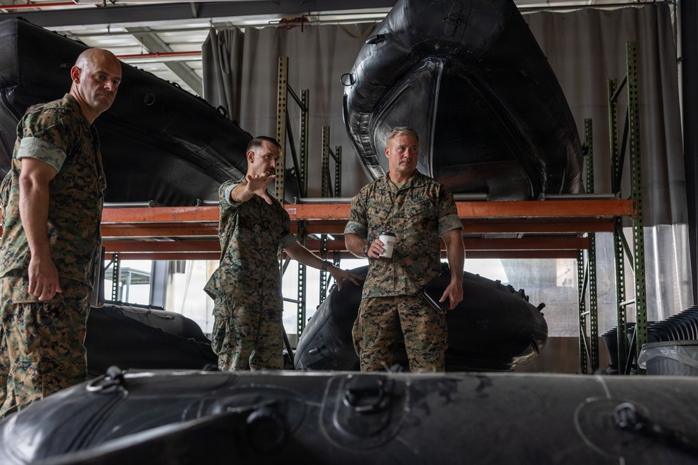 Three military personnel in MARPAT (Marine pattern) camouflage uniforms are standing in what appears to be a storage facility or warehouse. The central figure is gesturing and appears to be briefing or explaining something to the others, one of whom is holding a cup. The facility contains multiple large inflatable rubber boats (IBS - Inflatable Boat Small) stored on metal shelving racks.