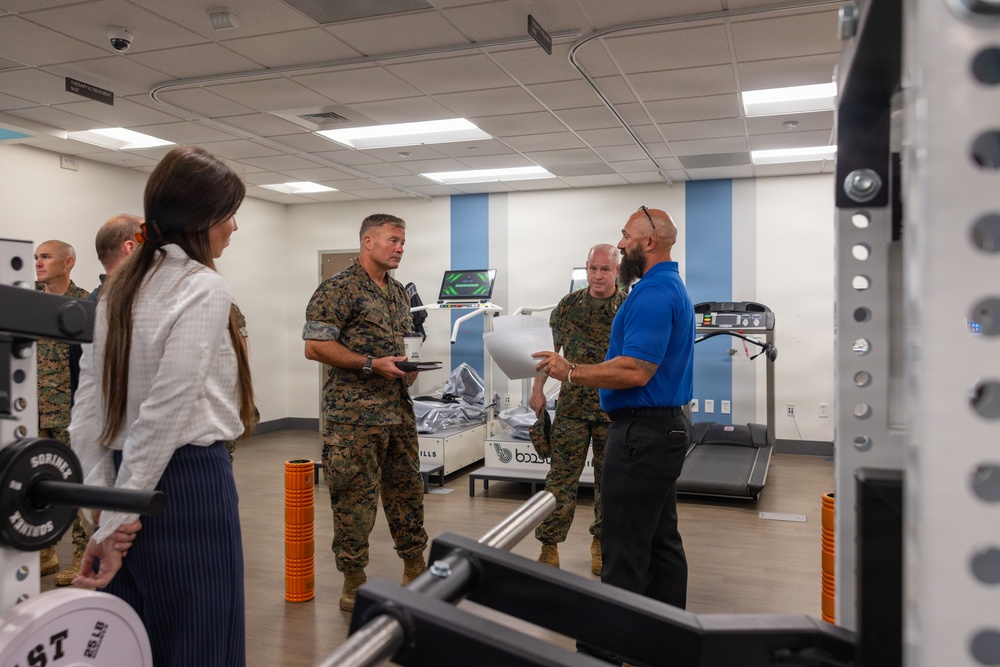 A group of U.S. Marine Corps personnel in camouflage uniforms are receiving a briefing or tour of a fitness/rehabilitation facility. A civilian in a blue polo shirt appears to be presenting information, holding documents, while speaking with the military personnel. The room contains exercise and rehabilitation equipment consistent with a military fitness or sports medicine center.