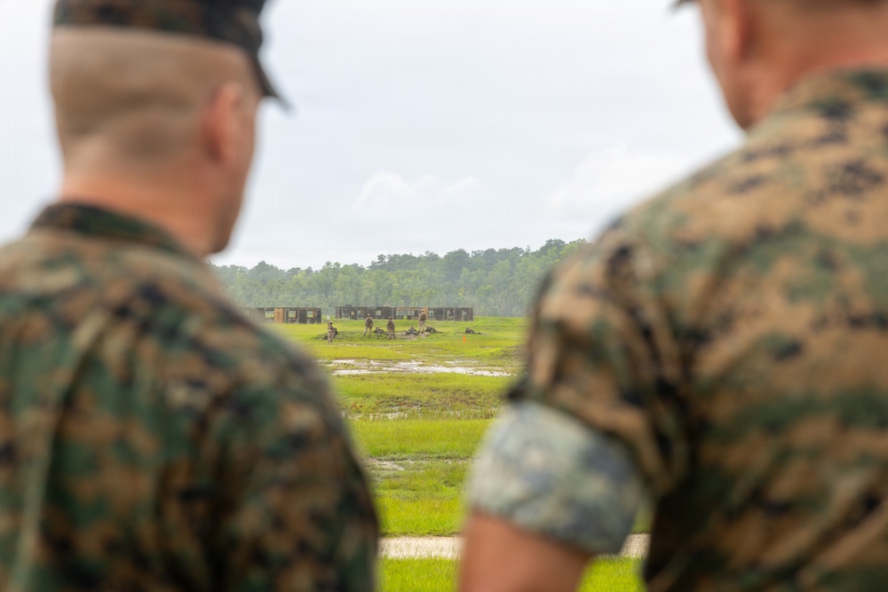 Two military personnel in digital camouflage uniforms are observed from behind, watching a distant training exercise on an open field range. In the background, several figures can be seen near what appear to be target structures or range facilities on a wet, grassy field. The scene suggests an outdoor military marksmanship or tactical training exercise being observed by supervisors or evaluators.