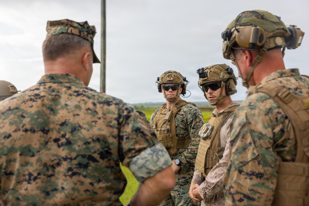 A group of military personnel, appearing to be U.S. Marines based on their MARPAT camouflage uniforms, are engaged in a briefing or discussion in an outdoor field environment. Several personnel are wearing tactical helmets with night-vision device mounts, body armor, and communication headsets, suggesting a training exercise or operational preparation. One individual facing away appears to be a senior officer or NCO holding a tablet or document.