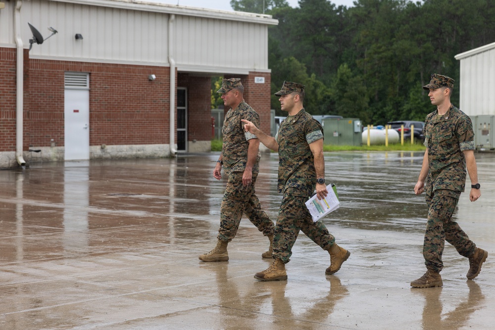 Three military personnel in MARPAT woodland camouflage uniforms are walking across a wet concrete surface during or after rainfall. The middle individual is carrying documents and appears to be briefing or conversing with the person to his left. The setting appears to be a military installation with brick buildings and a paved area.