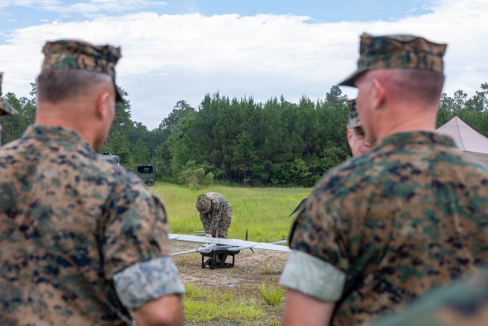Military personnel in woodland camouflage uniforms are gathered in an open field area observing a colleague preparing a fixed-wing unmanned aerial vehicle (UAV/drone) for operation. Two soldiers in the foreground watch as a third bends over the small aircraft on the ground. The setting appears to be a training or demonstration exercise at a military installation surrounded by pine trees.