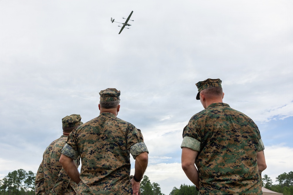 Three military personnel in MARPAT (Marine pattern) camouflage uniforms are observing a fixed-wing unmanned aerial vehicle (UAV) in flight overhead. The scene appears to be an outdoor demonstration or training exercise, likely at a military installation. The UAV visible in the sky has a distinctive twin-boom configuration consistent with reconnaissance drone designs.