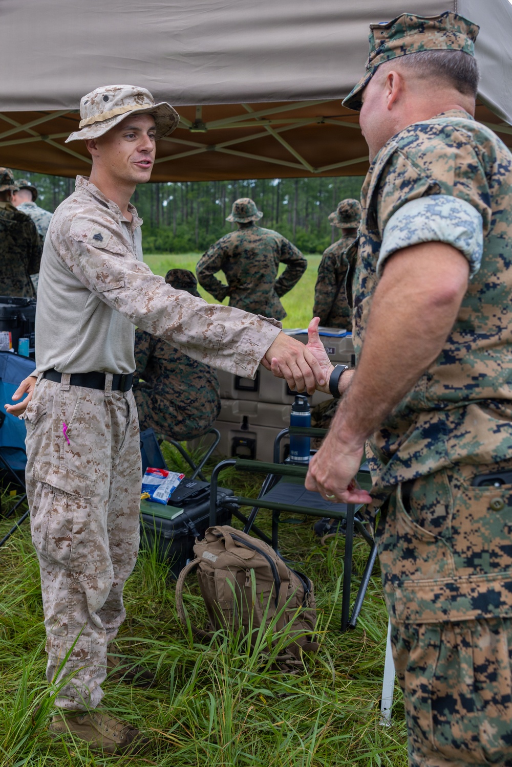 Two U.S. Marines in camouflage uniforms are shaking hands under a field canopy tent in an outdoor training or exercise environment. The Marine on the left wears desert MARPAT utilities with a boonie cover, while the one on the right wears woodland MARPAT with a cover. Several other Marines in the background appear to be observing an open field area.