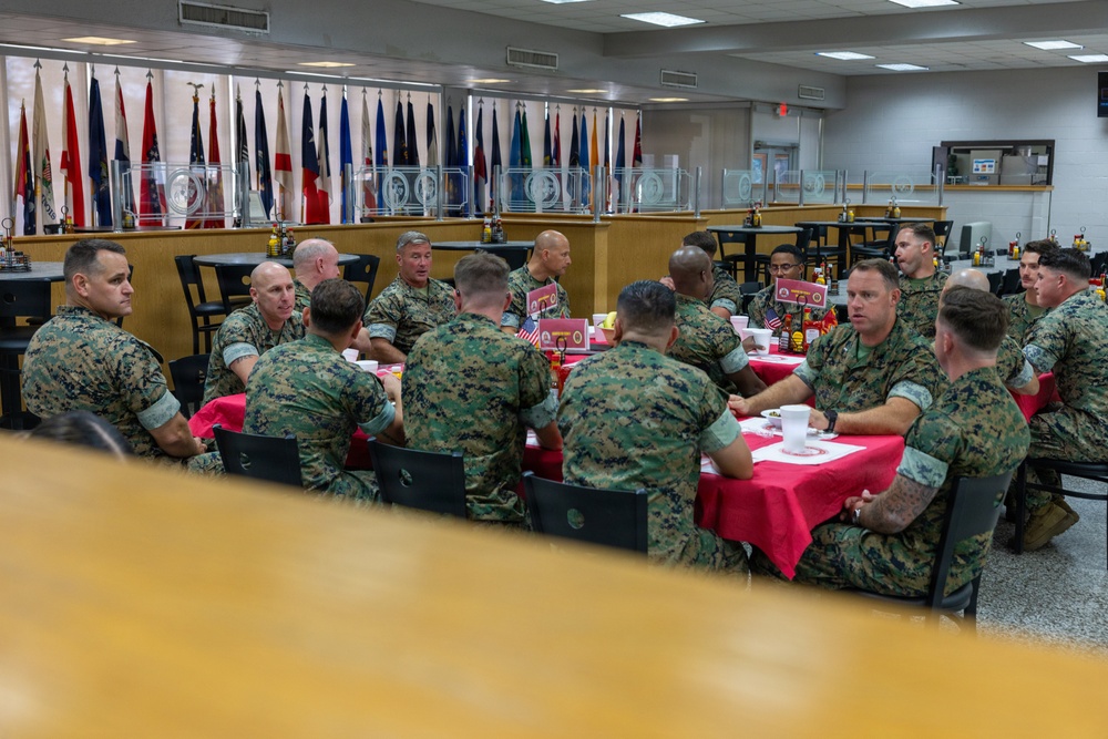 A group of U.S. Marine Corps personnel in MARPAT woodland camouflage uniforms are gathered around tables covered with red tablecloths in what appears to be a military dining facility or mess hall. The setting suggests a formal or semi-formal meeting, briefing, or unit gathering. Multiple state and military flags are displayed along the back wall, indicating an official military venue.