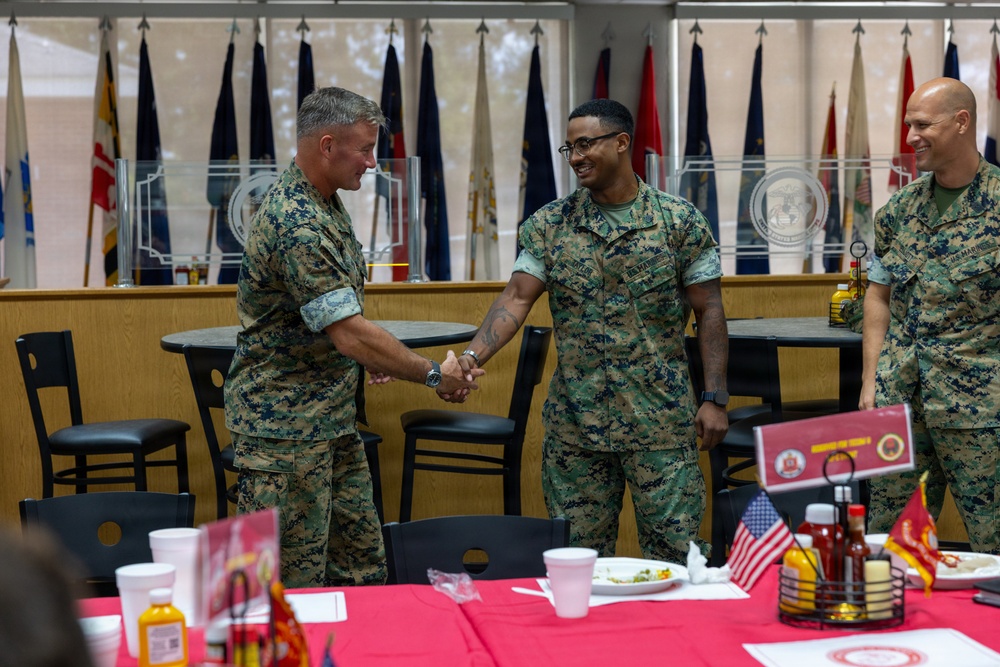 Three U.S. Marines in MARPAT camouflage uniforms are gathered in what appears to be a dining facility or mess hall. Two of the Marines are shaking hands in what appears to be a formal recognition or greeting ceremony, while a third Marine looks on smiling. The setting is decorated with multiple military unit flags and red tablecloths, suggesting a formal military dining event.