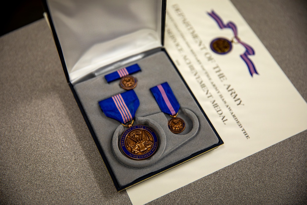 The image shows a Department of the Army Achievement Medal set displayed in an open presentation case, accompanied by an official award certificate. The set includes the full-size medal with blue and pink striped ribbon, a miniature medal, and a service ribbon bar. The items are arranged on gray velvet cushioning inside a navy blue clamshell box.