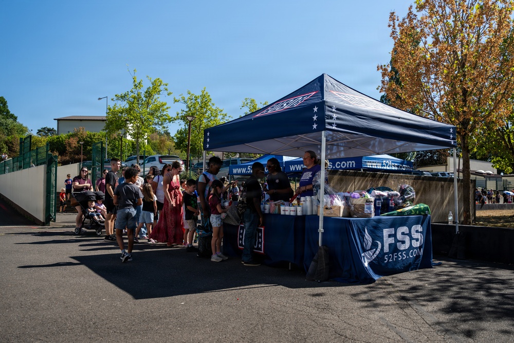 An outdoor community event or fair is taking place in a paved parking lot area on a sunny day. A navy blue canopy tent branded with 'FSS' and '52FSS.COM' serves as a booth where staff interact with a crowd of adults and children. The event appears to be associated with the 52nd Force Support Squadron, likely a U.S. Air Force installation community event.