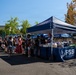An outdoor community event or fair is taking place in a paved parking lot area on a sunny day. A navy blue canopy tent branded with 'FSS' and '52FSS.COM' serves as a booth where staff are distributing or selling items to a line of attendees including adults and children. The event appears to be associated with the 52nd Force Support Squadron (52 FSS), a U.S. Air Force support organization.