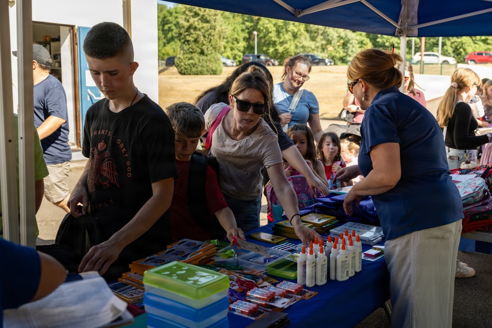 A community school supply giveaway event is taking place under a blue canopy tent outdoors. Families with children are gathered around a table covered with school supplies including glue bottles, pencil cases, folders, and other stationery items. A volunteer or organizer in a blue shirt is distributing supplies to attendees.