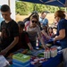A community school supply giveaway event is taking place under a blue canopy tent outdoors. Families with children are gathered around a table covered with school supplies including glue bottles, pencil cases, folders, and other stationery items. A volunteer or organizer in a blue shirt is distributing supplies to attendees.