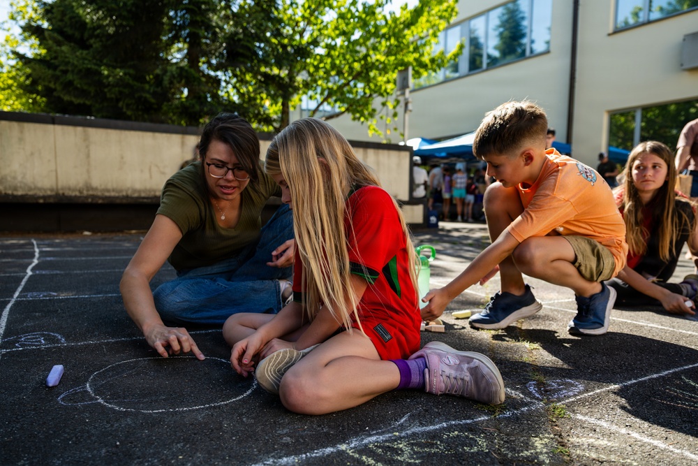 A group of children and an adult woman are engaged in sidewalk chalk drawing on an outdoor asphalt surface, likely a school or community courtyard. The adult, wearing glasses and a green shirt, is guiding the children in creating chalk artwork on the ground. The setting appears to be a warm, sunny day at an organized outdoor event or school activity.