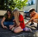A group of children and an adult woman are engaged in sidewalk chalk drawing on an outdoor asphalt surface, likely a school or community courtyard. The adult, wearing glasses and a green shirt, is guiding the children in creating chalk artwork on the ground. The setting appears to be a warm, sunny day at an organized outdoor event or school activity.